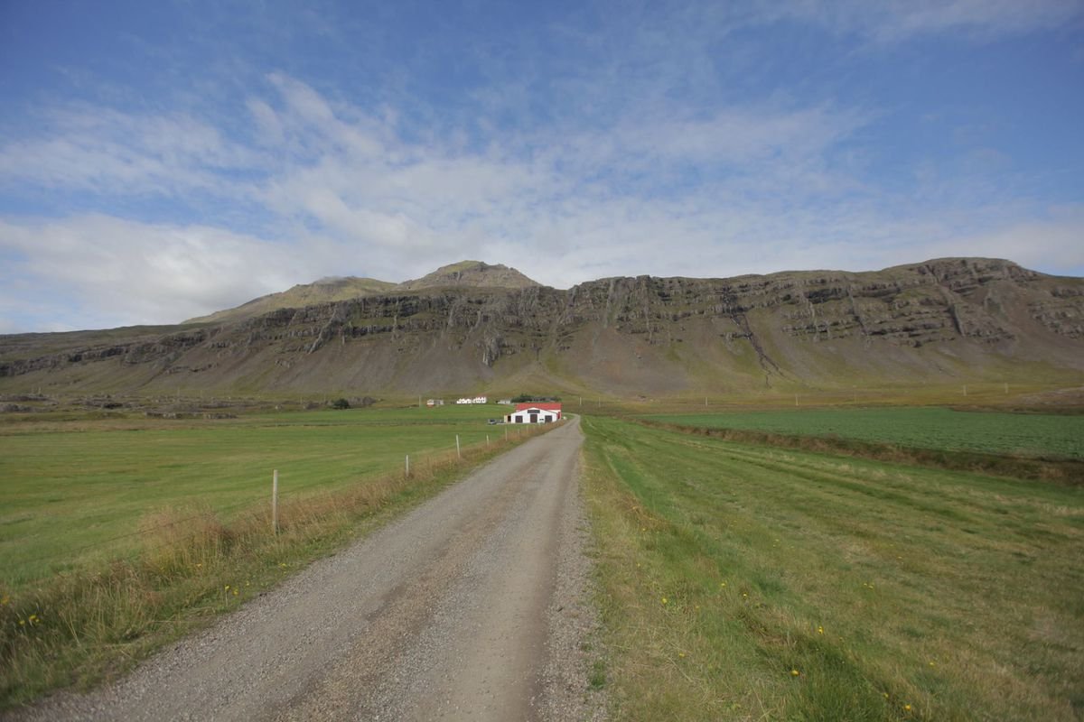 a dirt road with grass on both sides leading to a white barn with a red roof in the distance, sitting at the foot of some rocky hills