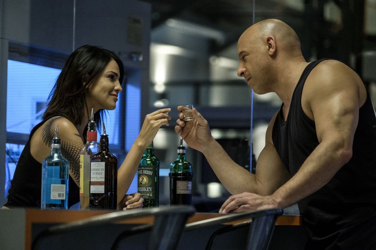Vin Diesel, in a black sleeveless T-shirt, clinks shotglasses with Eiza Gonzalez, a dark-haired woman with an elaborate black tattoo of scales running down her left arm, in Bloodshot.