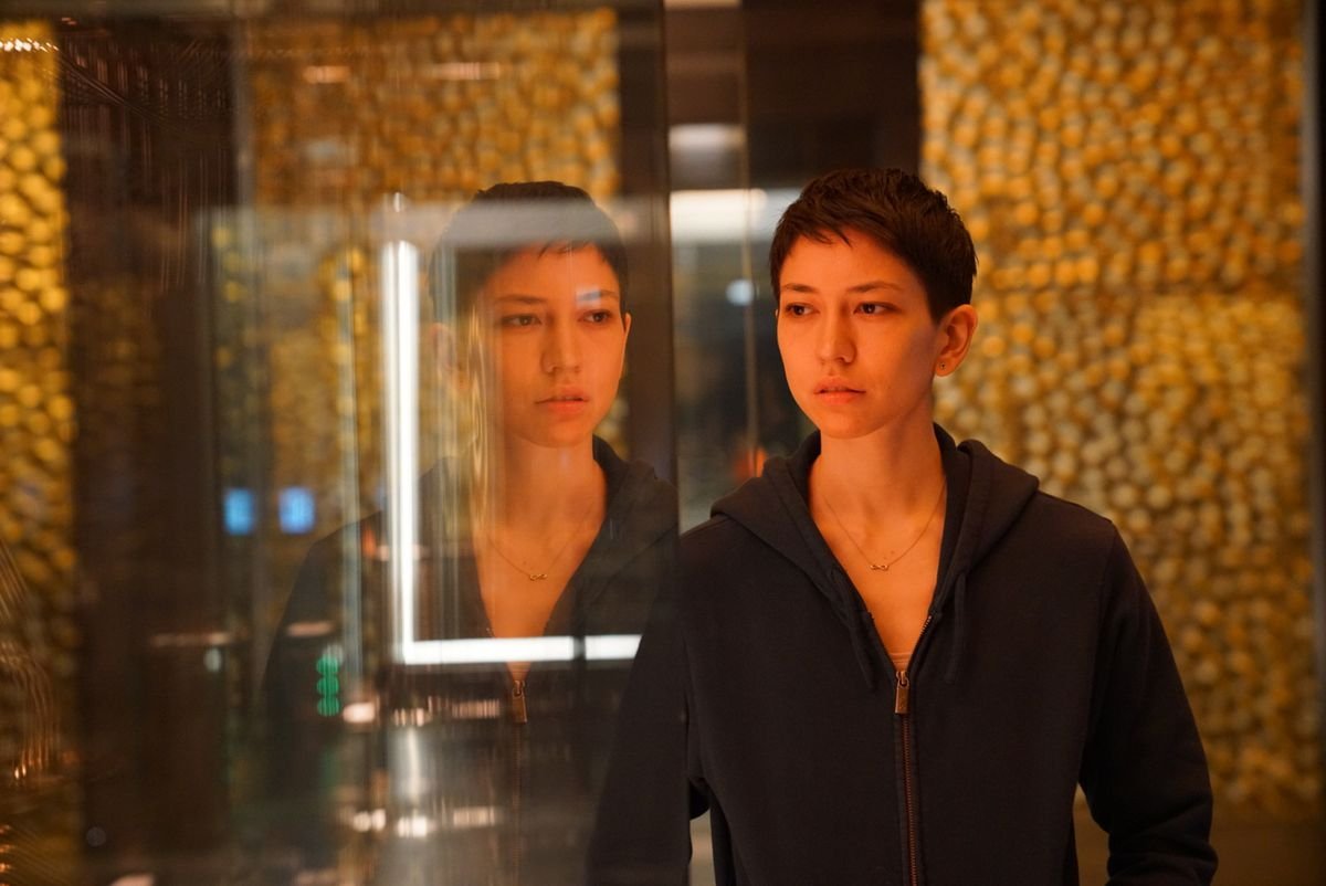 A short-haired Asian woman in a loose-fitting black hoodie stands in front of a glass wall staring outward, as the reflection behind her appears to stare over her shoulder.