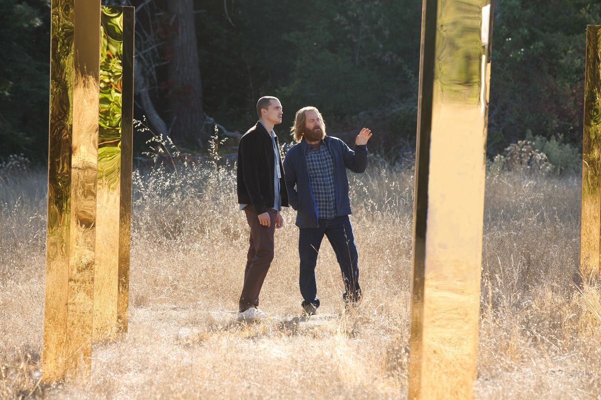 Parks and Recreation’s Nick Offerman, with long hair and a heavy beard, stands in a pale, grassy field with a forest backdrop, amid tall, reflective golden metal columns. He has a hand up, gesturing toward one of the columns, and appears to be explaining something to a thinner, more clean-cut, younger male associate.