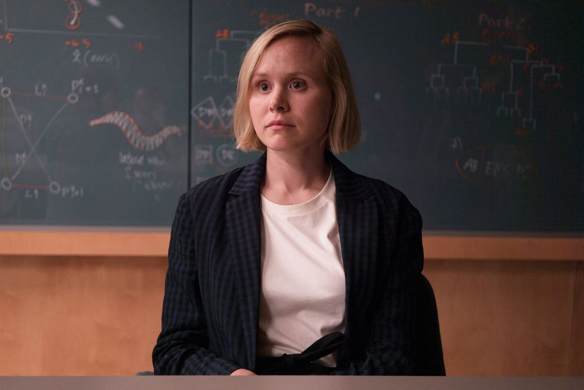 A woman with short blonde hair and a fixed, wide-eyed expression sits staring off-camera in front of a chalkboard covered with diagrams that appear to be programming branch trees and other configurations.