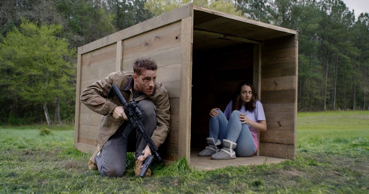 A scruffy-looking, bloody-faced man crouches behind a huge wooden crate and clings to a handgun and a rifle, while a frightened-looking woman in pastels sits inside the crate, staring in his direction.