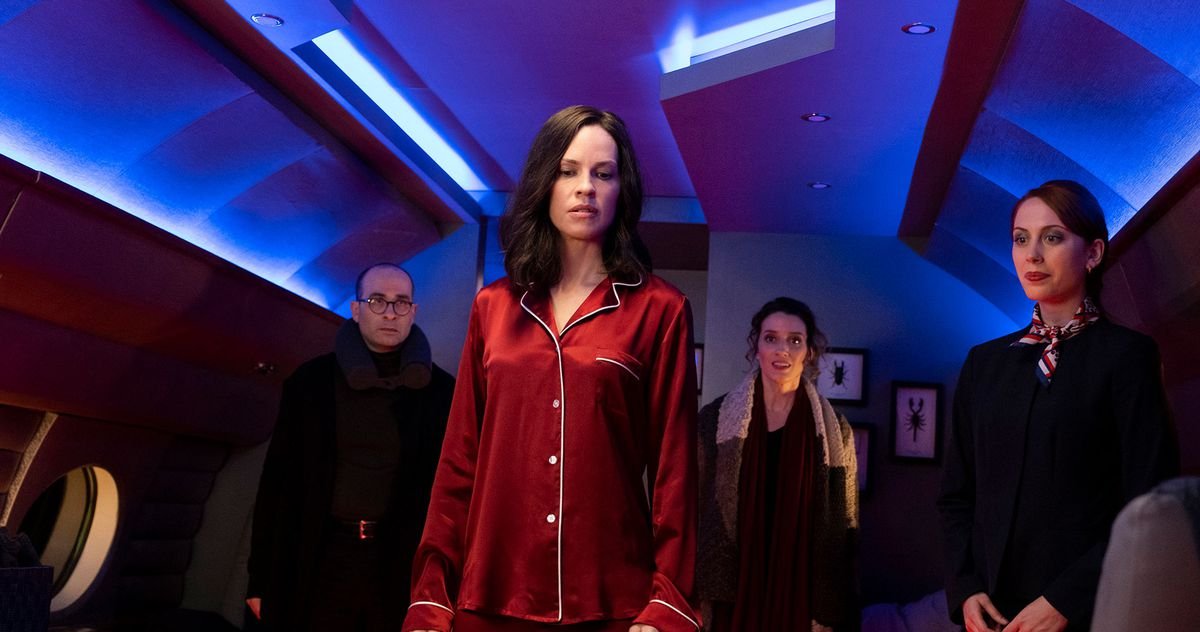 Hilary Swank, in red pajamas, looks disdainfully toward something below the level of the screen, in a blue-lit space with framed scorpions and beetles on the wall, as three fully dressed people circle her.