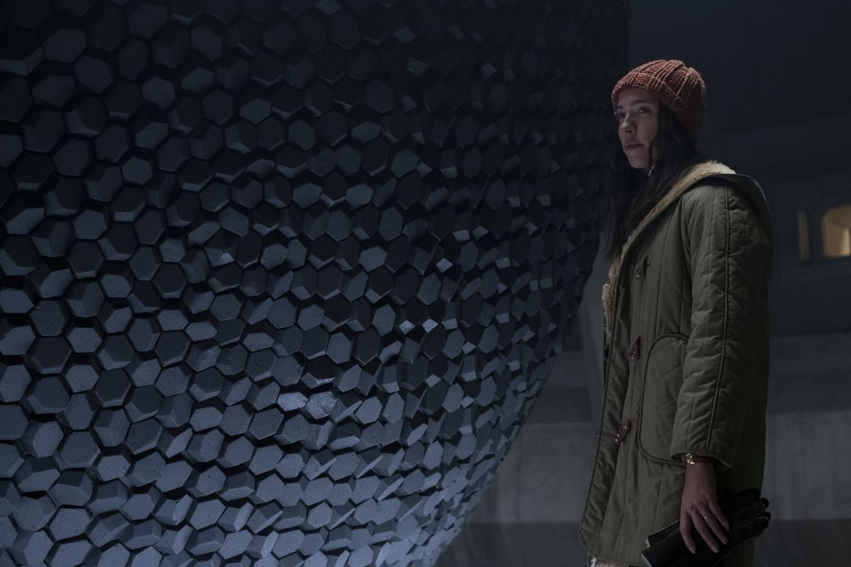 A woman, the daughter of the director and the inheritor of the lab upon his death, stands in front of an amorphous sphere made of dark gray stones.