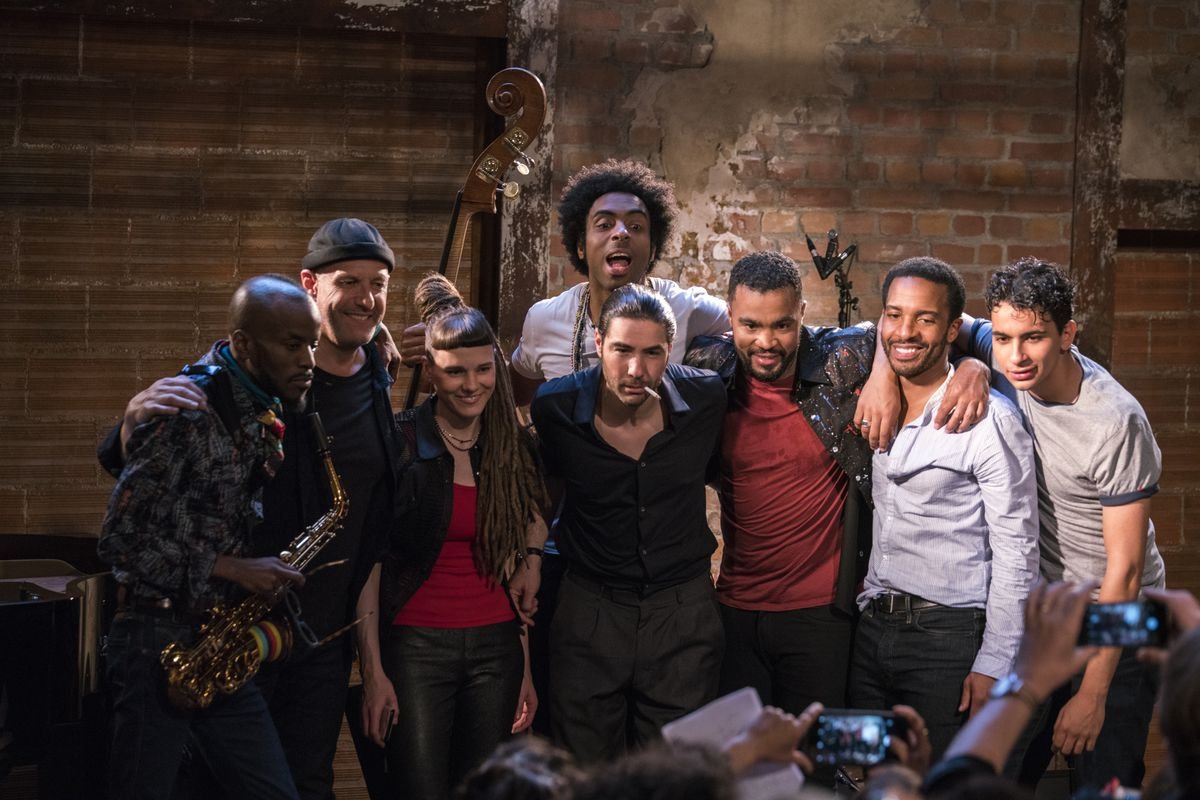 The eight-person band from The Eddy poses together, arms draped over each over, smiling at a group of people holding up their phones to take photos of the group.