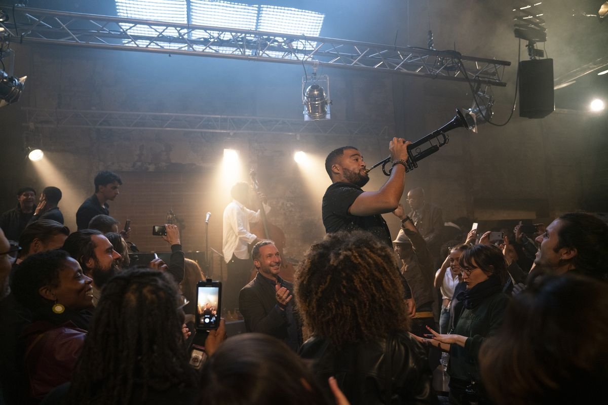 In a scene from The Eddy, an audience gathers around a performer who’s standing on stage, blowing into a muted black trumpet.