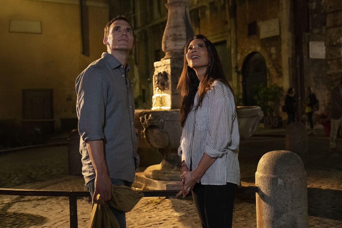 Sam Claflin and Olivia Munn, dressed as basically as humanly possible, stand outside in front of a Roman fountain, both staring upward and offscreen as though there’s something interesting above them.
