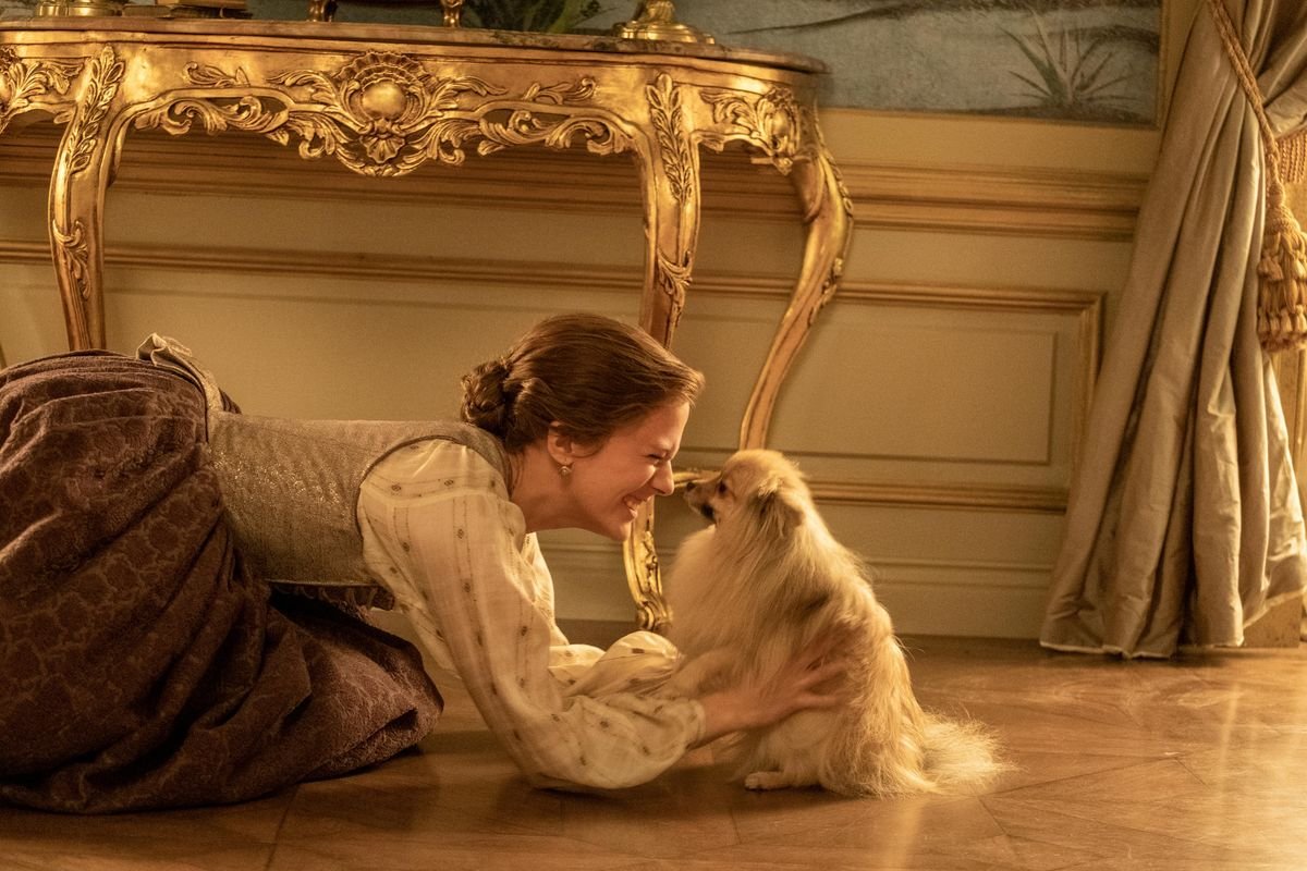 A woman on her knees in front of an elaborate gold-leafed table nuzzles a small fuzzy dog in Hulu’s historical drama The Great.