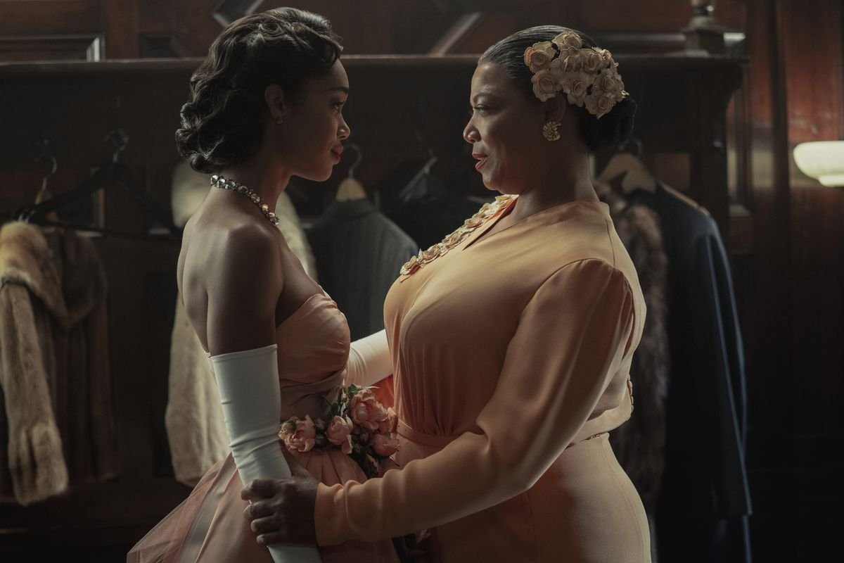 An older black woman in a conservative peach dress speaks to a younger black woman in a bridesmaid’s-style peach dress in front of a group of hanging coats.