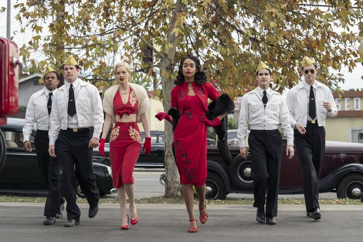 A white woman and a black woman in form-fitting red outfits cross a parking lot, in front of four men in white jackets, black ties, and peaked khaki garrison hats.