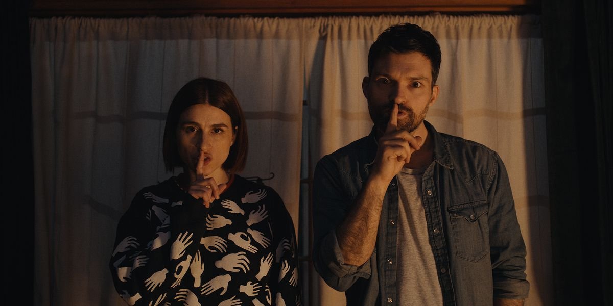 Josh Ruben and Aya Cash stare into the camera and make the “shhh” gesture in Scare Me