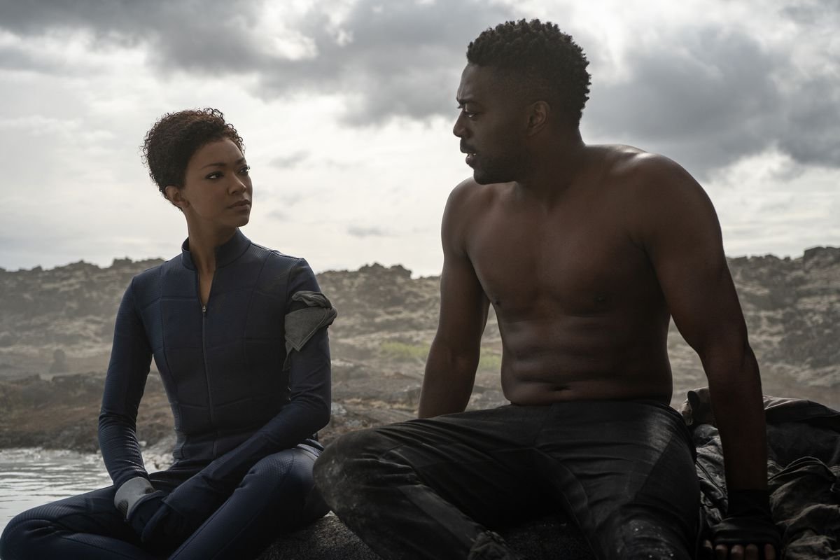 Sonequa Martin-Green sits next to shirtless David Ajala in a barren landscape by water in Star Trek: Discovery