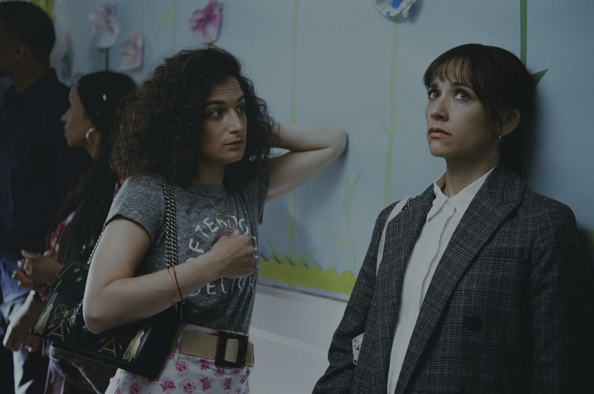 Jenny Slate, looking curious, leans against a wall with Rashida Jones, looking exasperated, in On the Rocks