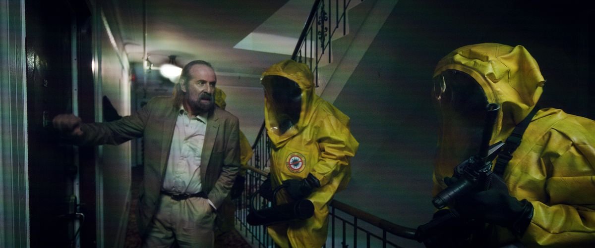 Peter Stormare points two people in yellow HAZMAT suits at an apartment door in Songbird
