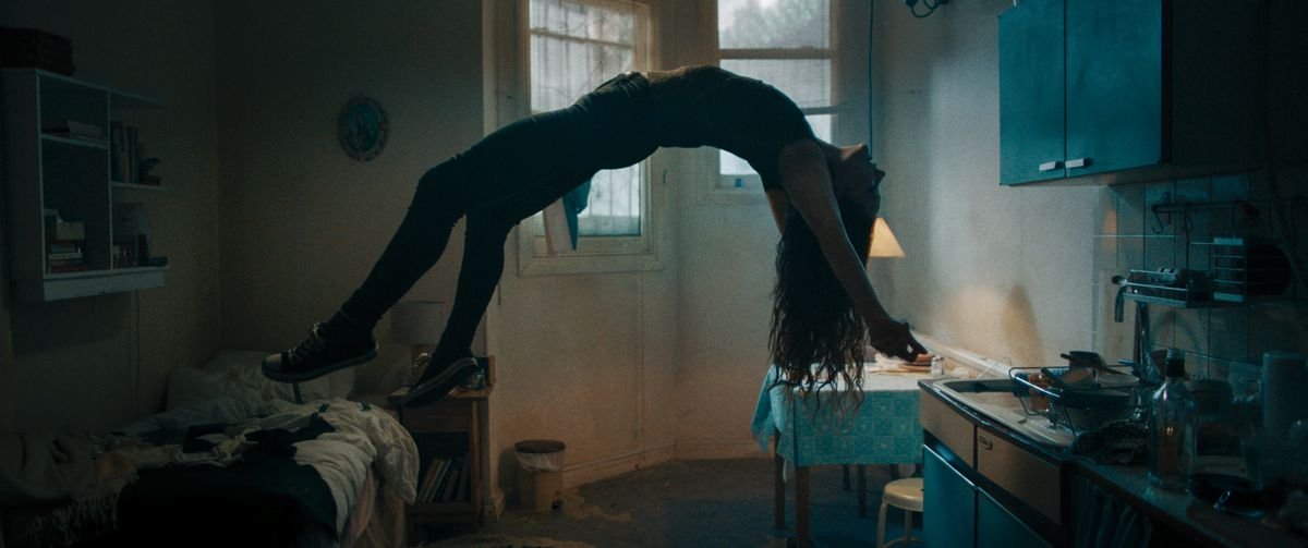 A woman floats on her back in mid-air in a dark room, back arched and long hair dangling