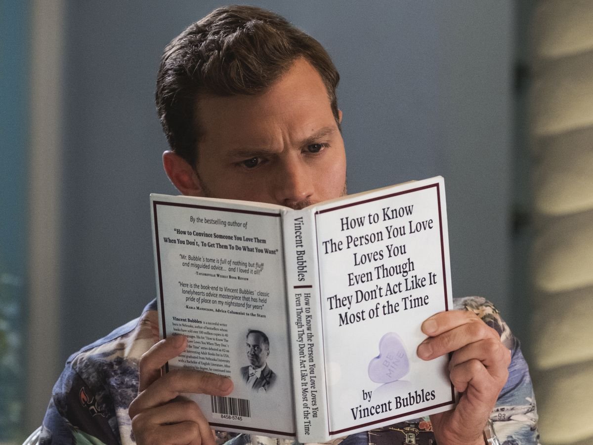 Jamie Dornan as Edgar reading a book called How to Know the Person You Love Loves You Even Though They Don’t Act Like It Most of the Time in Barb & Star Go to Vista Del Mar