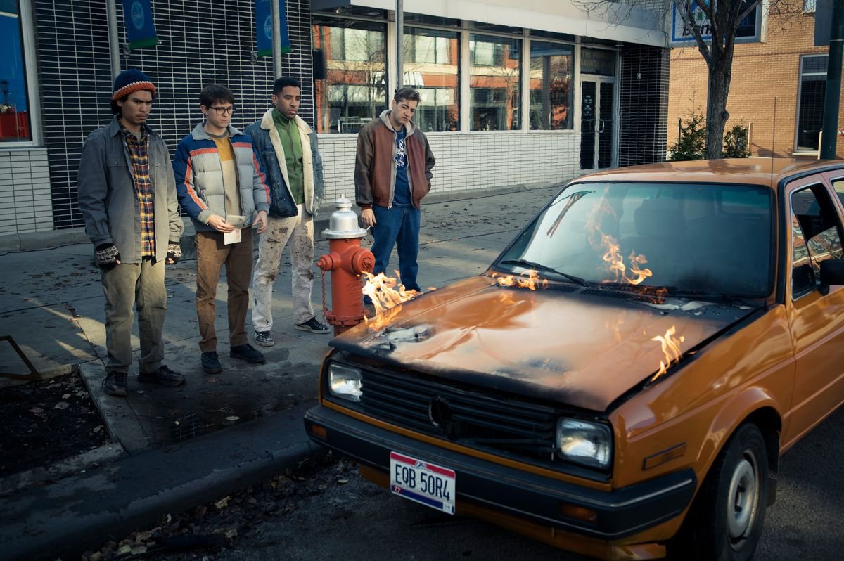 Cherry and his crew stand next to an orange car as the hood catches on fire