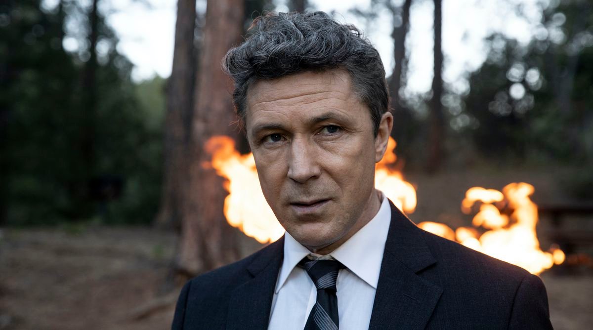 Aidan Gillen in the woods with a fire behind him in Those Who Wish Me Dead