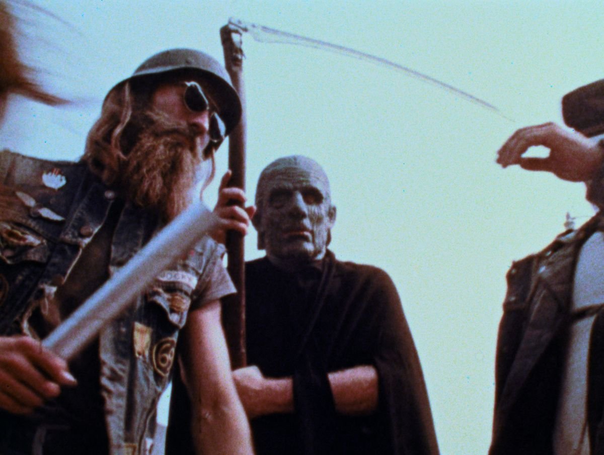 A thuggish-looking biker-type wielding what looks suspiciously like a silver paper-towel tube and a bald man in black with a scythe looking like the Grim Reaper in George A. Romero’s The Amusement Park