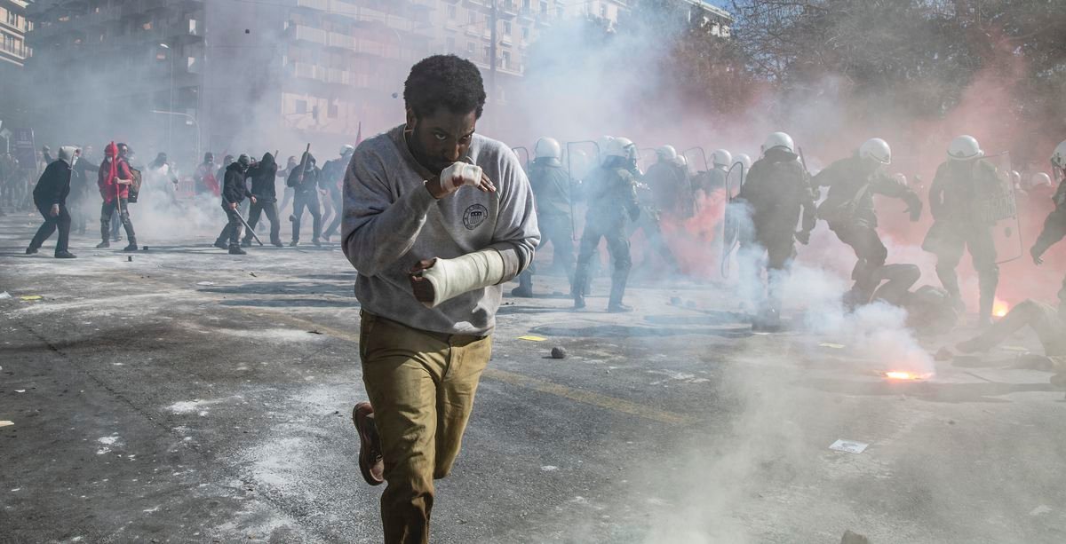 Beckett protagonist John David Washington, hands bandaged and bloodied, runs through the smoke-filled streets as a riot takes place in the background