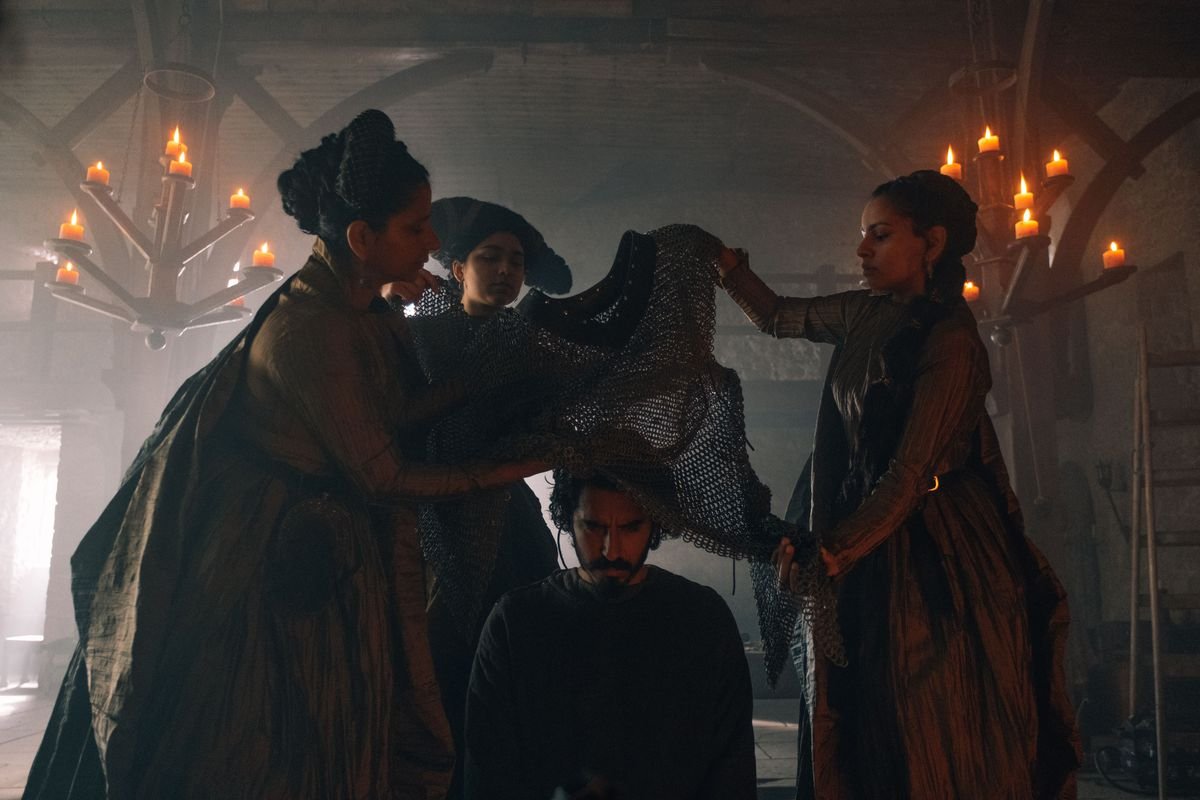Three women lower a chain shirt over a kneeling Dev Patel in David Lowery’s The Green Knight