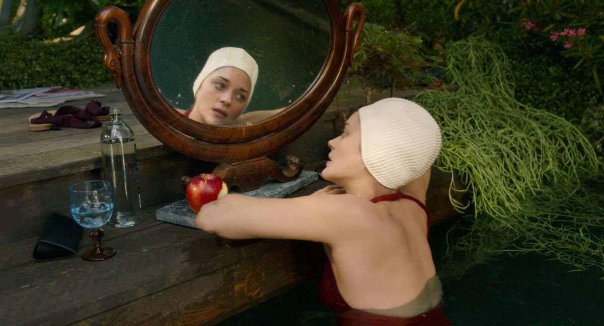 Marion Cotillard, in bathing suit and white swim cap, examines herself in a round mirror in Annette
