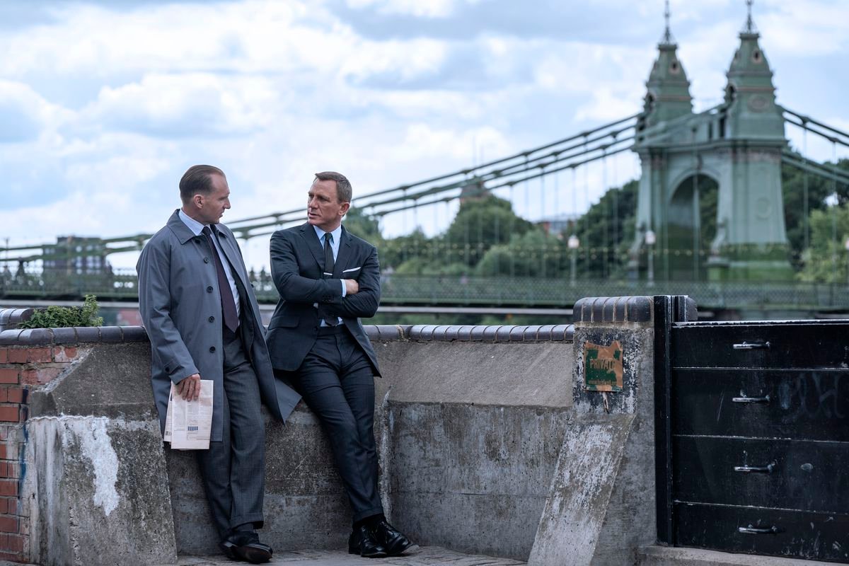 Daniel Craig as James Bond and Ralph Fiennes as M enjoy a leisurely outdoor meeting near a bridge in No Time To Die