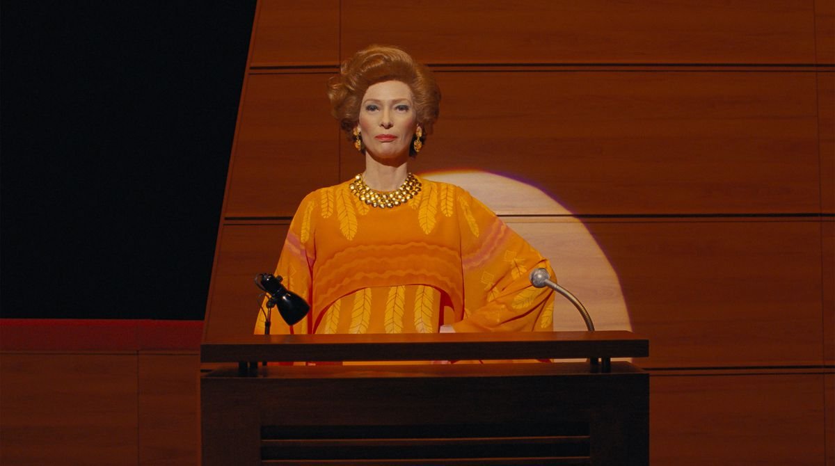 Tilda Swinton, in an orange bouffant and blaze-orange layered dress, at a spotlighted podium in Wes Anderson’s The French Dispatch