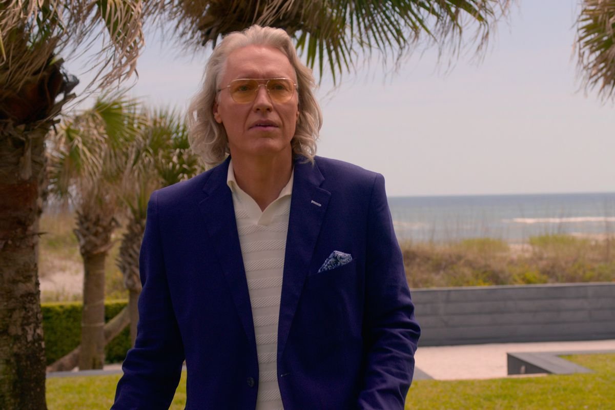 A man with tinted, wire-rimmed glasses and long, gray hair wearing a blue sports jacket. The beach is in the distance.
