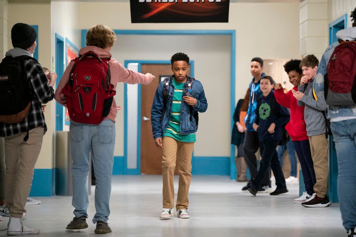 A junior-high age boy with a backpack over one shoulder walks alone in the hall as someone points at him