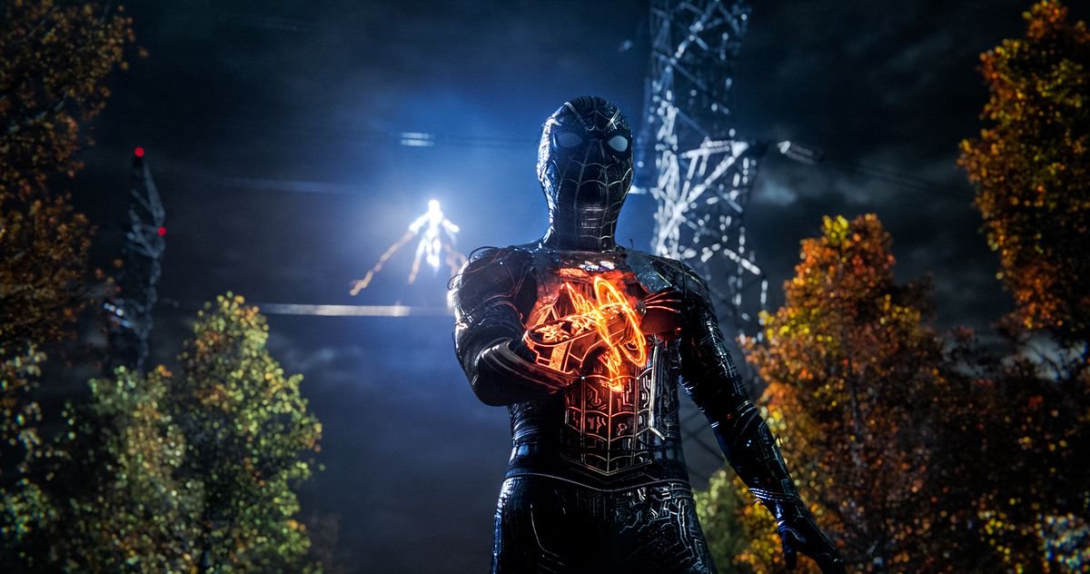 Spider-Man, in a black suit with a mystical device on his wrist, stands in a dark woods while Electro appears in the background in Spider-Man: No Way Home.
