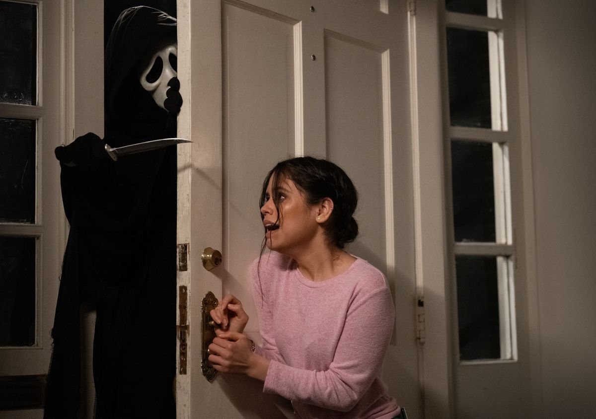 Jenna Ortega leans against a door, screaming, as Ghostface tries to burst in in 2022’s Scream