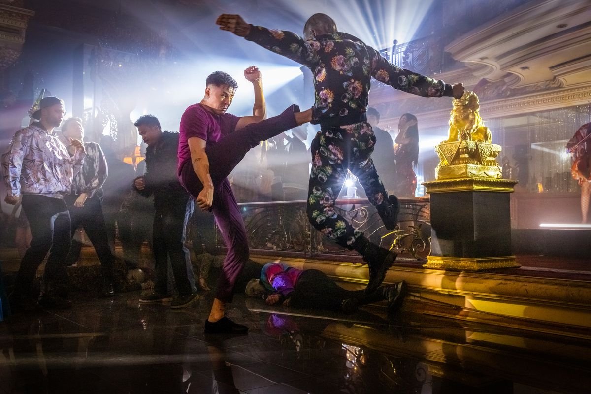 Lewis Tan, in loose purple pants and shirts, kicks a flying weirdly pajamaed man in Fistful of Vengeance