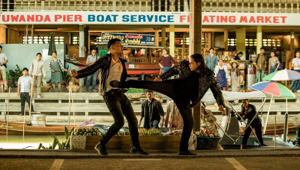 Lewis Tan, in all black with a black-and-gold jacket, totally kicks some dude right in the stomach in an open-air market in Fistful of Vengeance