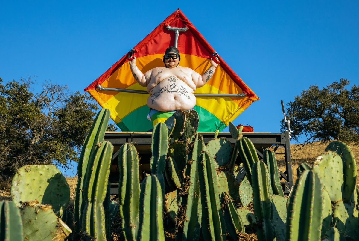 Zach Holmes, an extremely large and shirtless tattooed man, holds up a hang glider and smiles as he stands above a field of cacti
