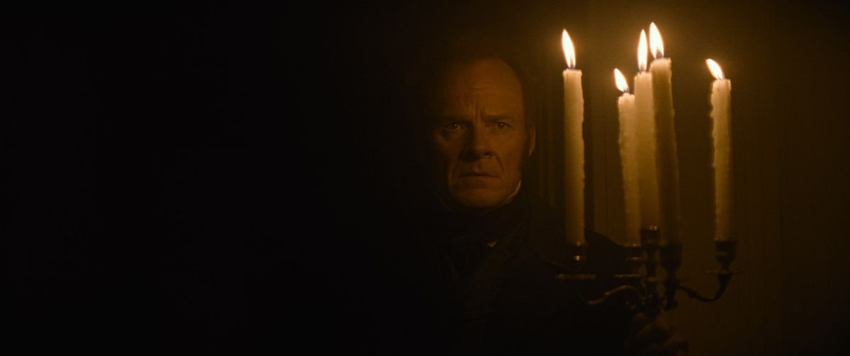 Alistair Petrie in an incredibly dark room in The Cursed, lit only by the candelabra he’s holding