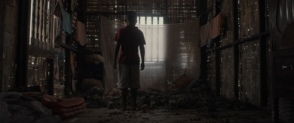 A young Lao boy stands with his back to the camera, looking at a pile of detritus in a dark, cluttered room in Mattie Do’s The Long Walk