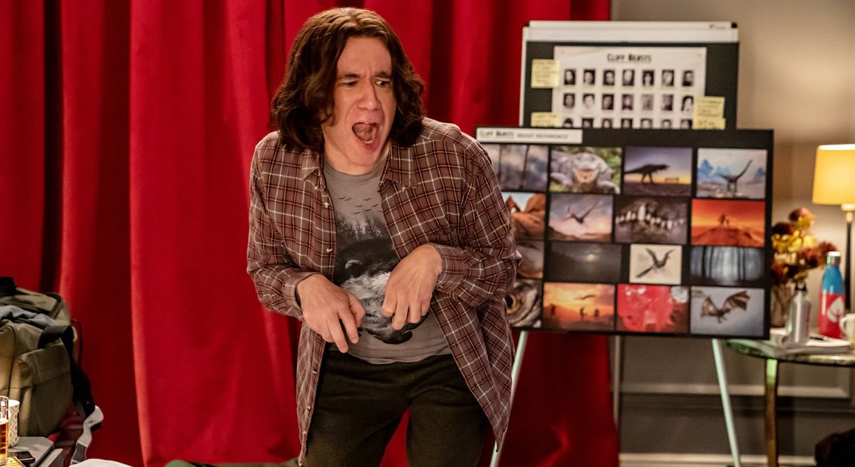 Fred Armisen in a shoulder-length curly wig stands in front of a mood board of dinosaur images with his hands drawn up against his chest like T Rex. arms and roars in The Bubble