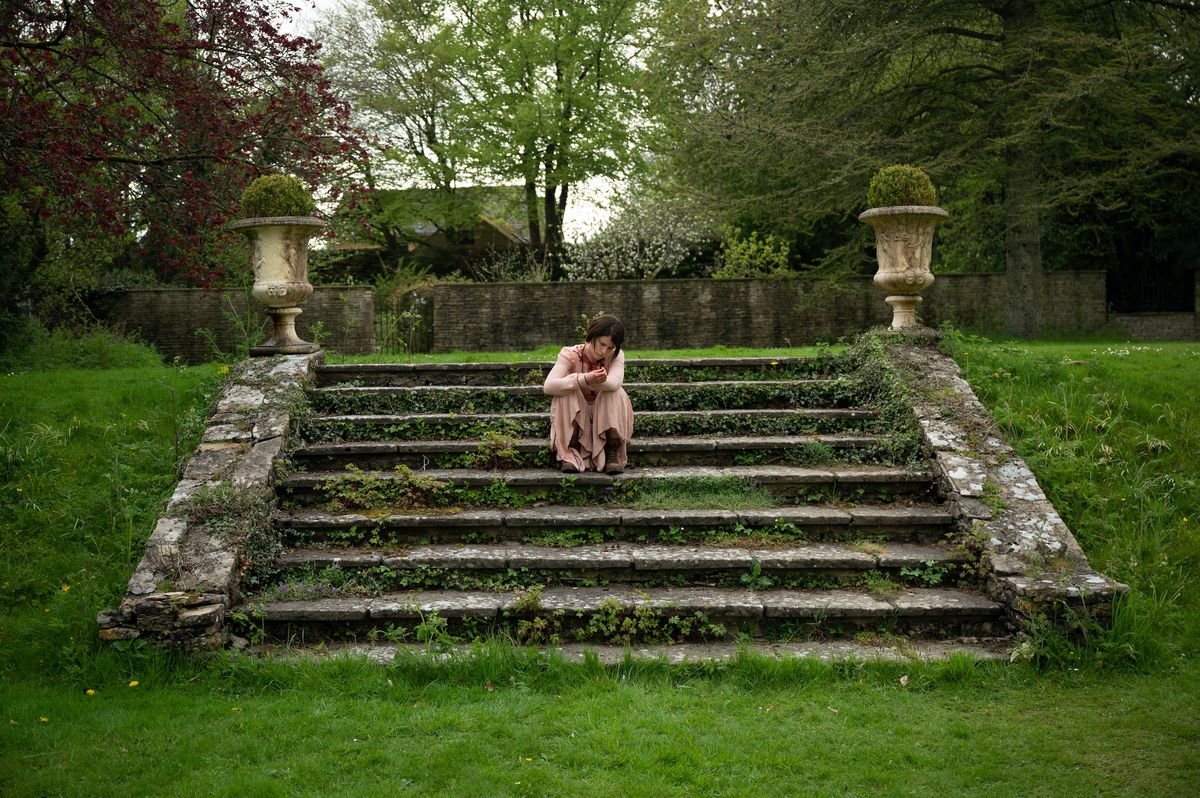 Jessie Buckley, in a long pink dress, sits on moss-covered stairs in a green landscape in Alex Garland’s Men.