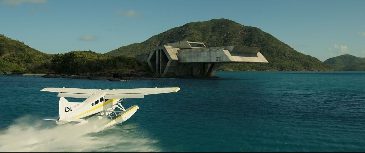 A white seaplane against a vivid dark-blue ocean approaches the Spiderhead prison, a stylized concrete building jutting out of an island like a fortress