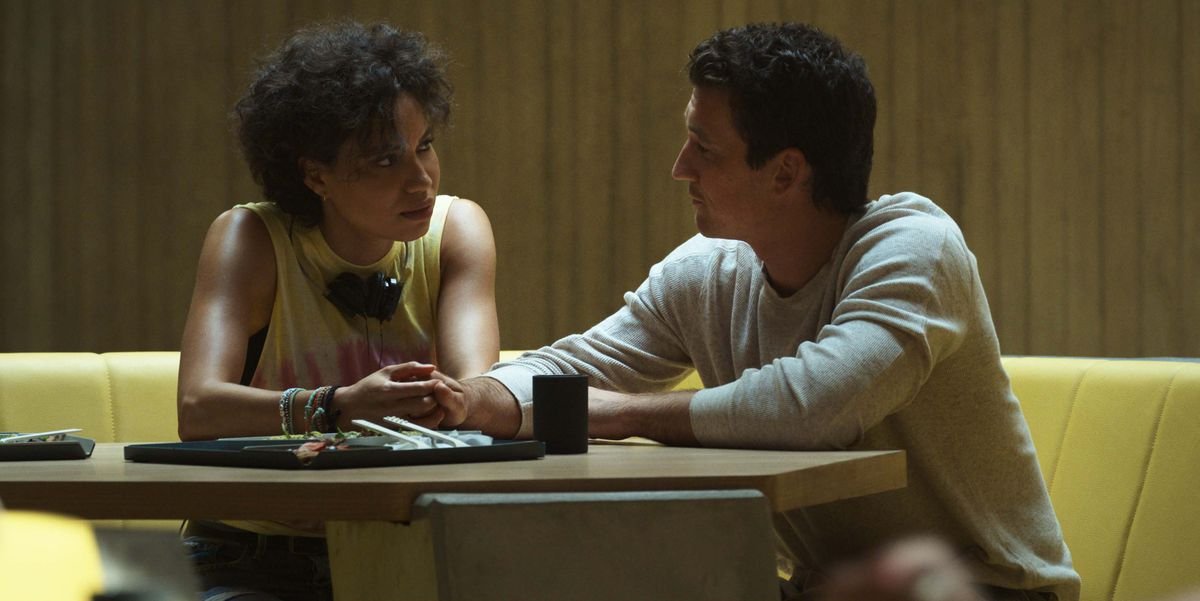 Jurnee Smollett and Miles Teller sit together at a table, touching hands, in the Spiderhead prison in Netflix’s Spiderhead