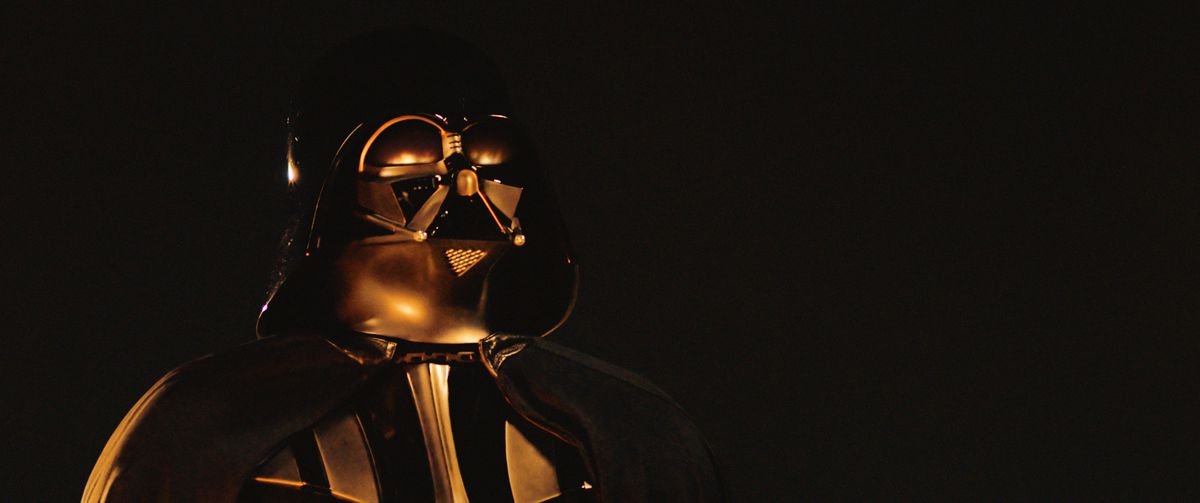 Darth Vader stands in the darkness as flames reflect in his suit