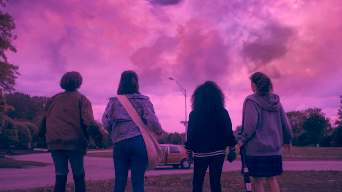 The four paper girls standing and facing away from the camera at the bright pink sky