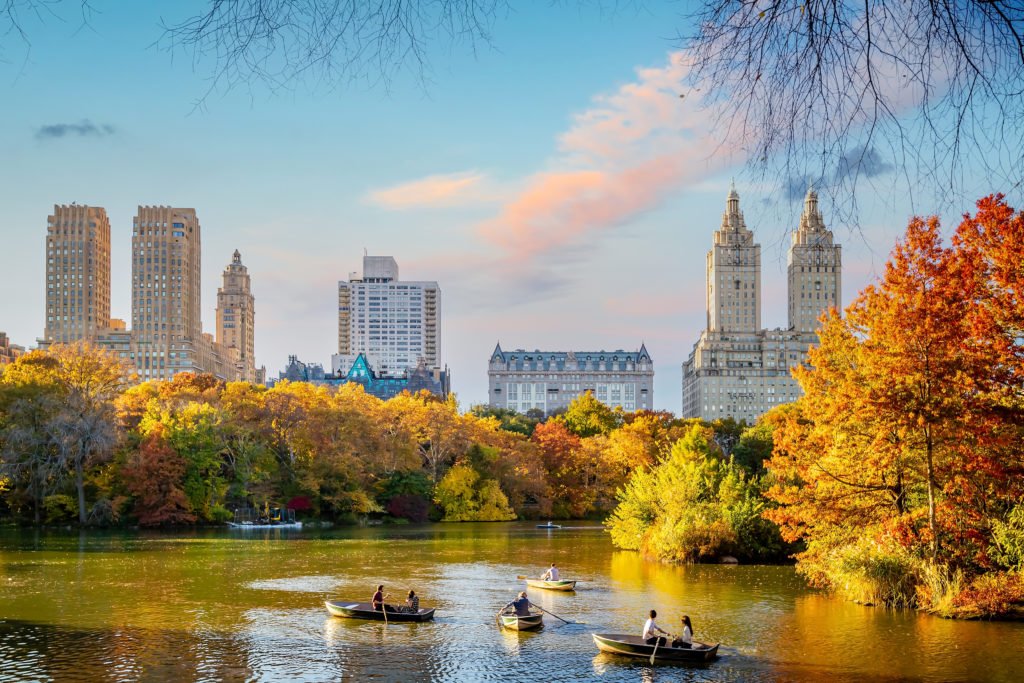 People paddling boats on the water in Central Park surrounded by autumn leaves and the New York City skyline in the background