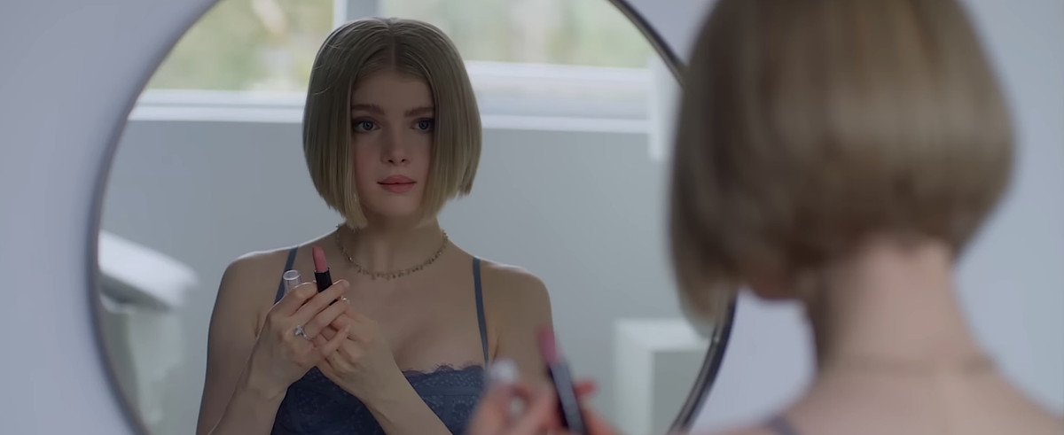 Meredith (Elena Kampouris) examines herself in a mirror while holding up lipstick in Wifelike