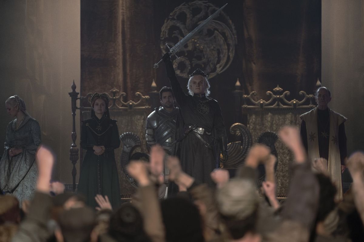 King Aegon II (Tom Glynn-Carney) standing in front of a crowd of cheering Westeros members of the public, holding his sword in the air triumphantly 