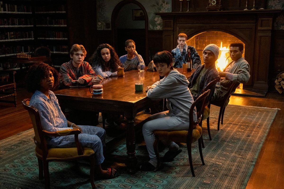 The members of the Midnight Club sitting around a table in the library looking towards a camera. There’s a fireplace with a fire behind them