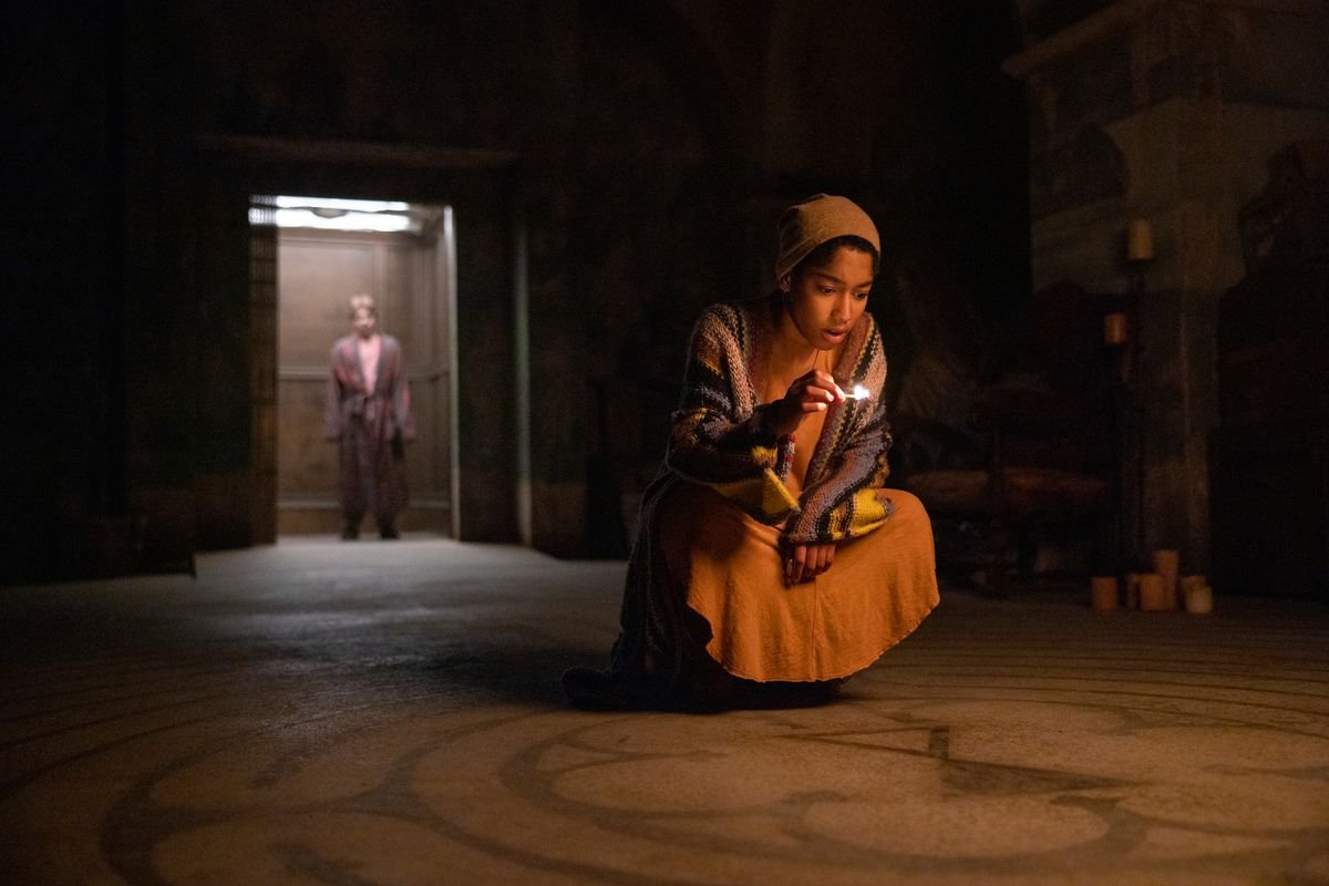 Ilonka kneeling with a friend in the lit elevator behind her. She is holding a match and looking at a pattern on the ground of a dark basement room.