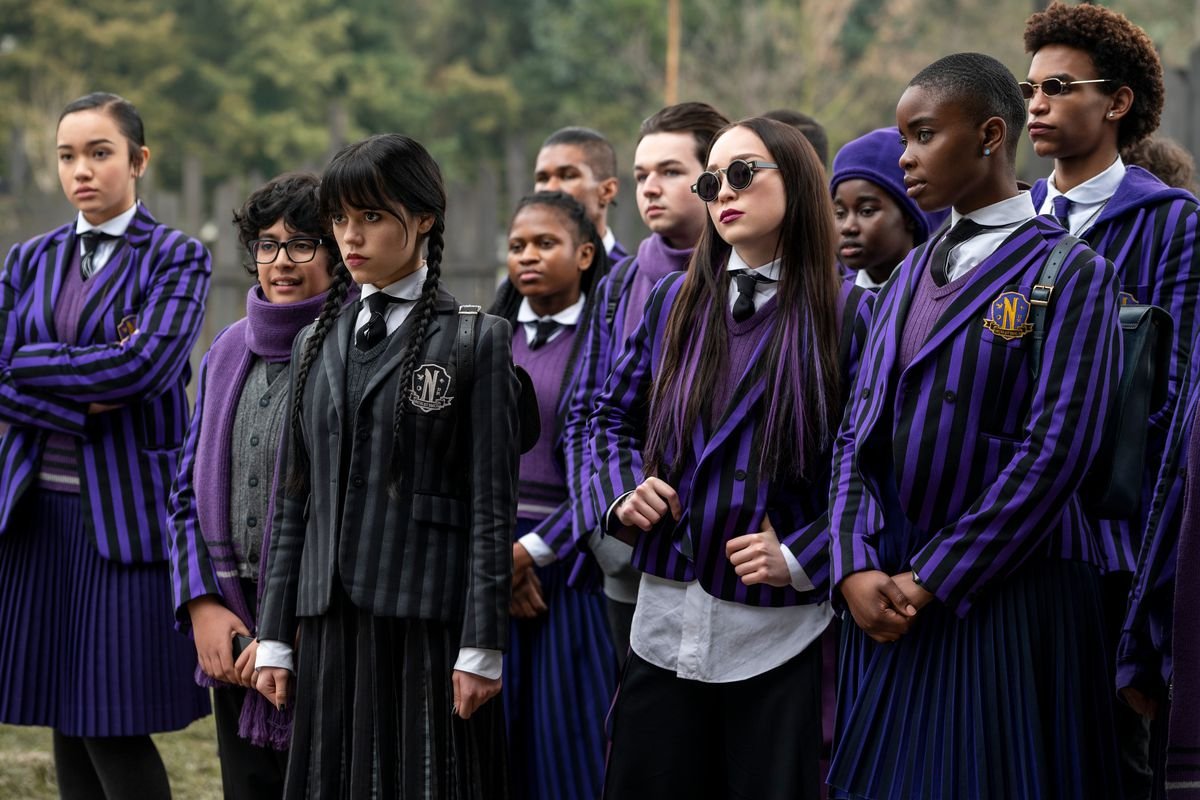wednesday stands with a group of students from nevermore academy; they all wear dark blue pinstripe uniforms, except for wednesday who wears black