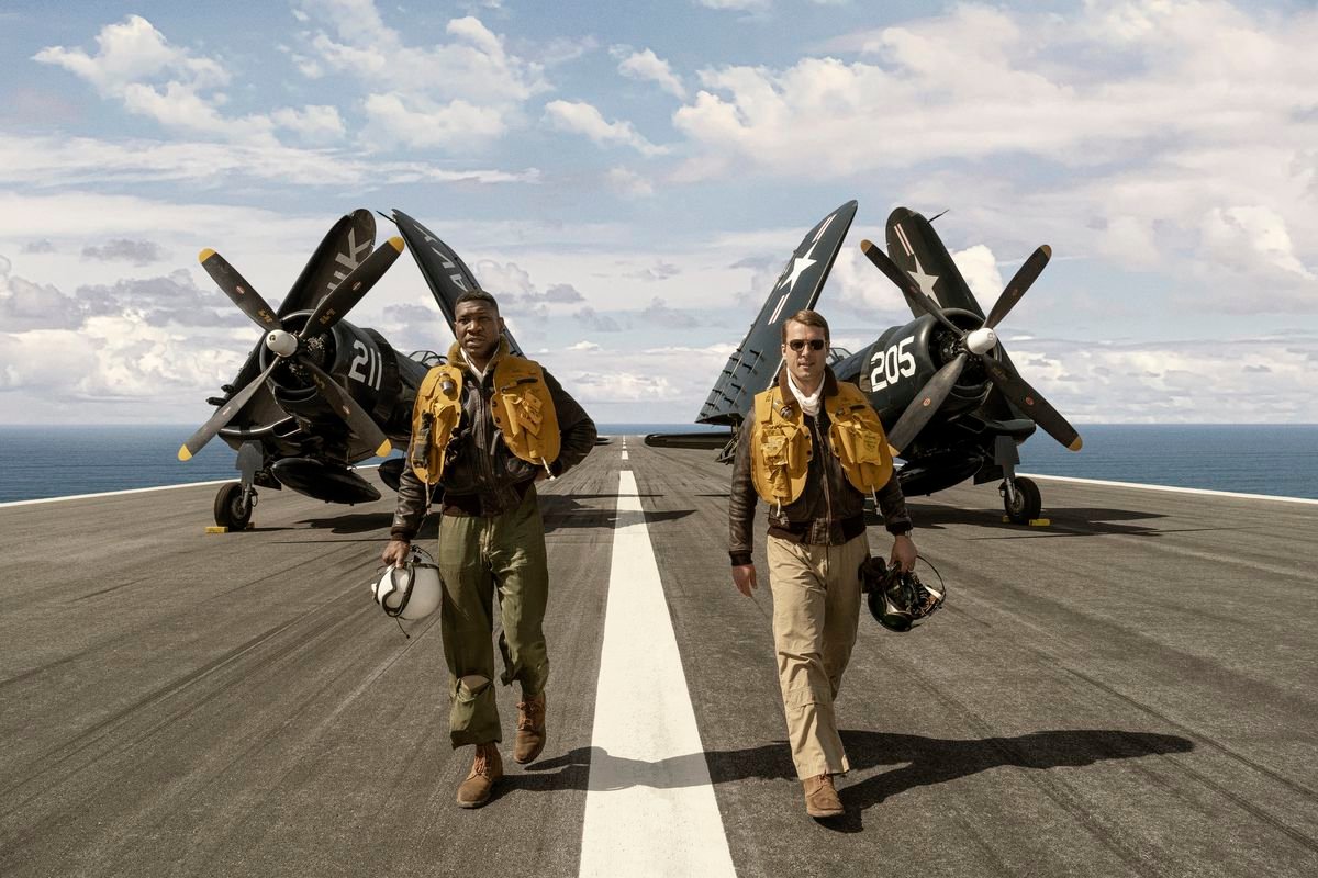 Jesse Brown (Jonathan Majors) and Tom Hudner (Glen Powell), in Navy pilot gear, walk toward the camera with their planes in the background on the deck of an aircraft carrier with the sea visible beyond it in Devotion